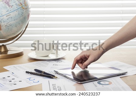 A working wooden desk(table) with tablet pc, coffee cup, graph, globe, paper, pencil and hand behind white blind(roller blind, shade).