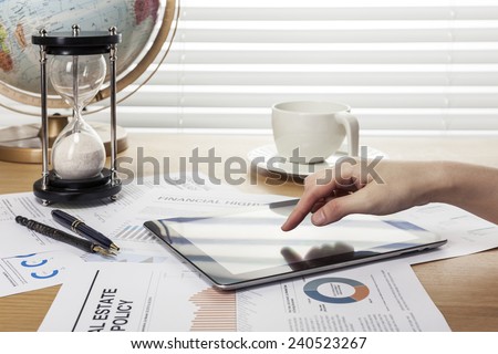 A working wooden desk(table) with tablet pc, coffee cup, graph, globe, sand timer, paper, pencil and hand behind white blind(roller blind, shade).
