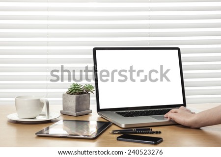 A working wooden desk(table) with notebook computer, tablet pc, mobile, hand phone, coffee cup, globe, pencil, behind white blind(roller blind) and hand.