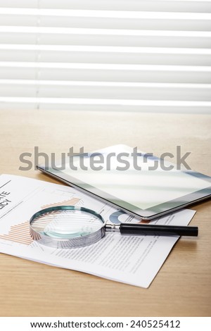 A wood working(office) table(desk) with a magnifier(reading glasses, magnifying glass[lens]), tablet pc  on the graph paper(document)s for business behind blind(rolling blind, shade).