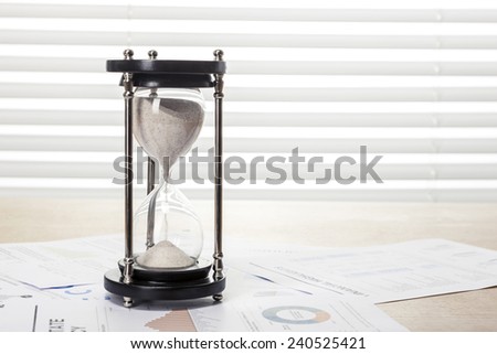 A wood working(office) table(desk) with sand timer(hour glass) on the graph paper(document)s for business behind blind(rolling blind, shade).
