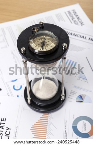 A wood working(office) table(desk) with sand timer(hour glass), graph paper(document)s for business, vintage compass, top view.