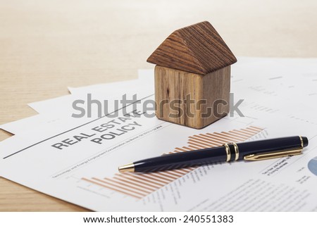 A house(made in wood block) with pen and graph paper(document) on the wood office desk(table) behind white blind.