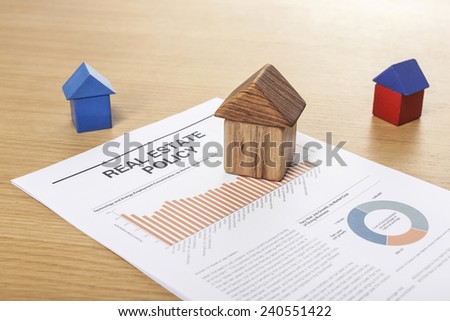 Three houses(made in wood block, red and blue) with graph paper(documents) on the office desk(table) behind white blind.
