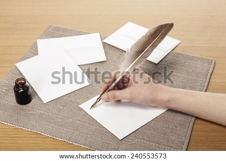 A female(woman) hand hold(write) a vintage feather quill pen with envelope and letter, ink on the office desk(table) behind white blind