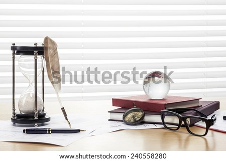 A sand timer(hour glass), feather quill pen, glasses, glass globe, compass, graph paper(document) and book on the wooden office desk(table) behind white blind.