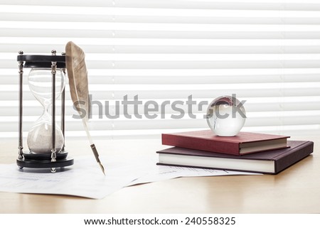 A sand timer(hour glass), feather quill pen, glass globe, graph paper(document) and book on the wooden office desk(table) behind white blind.