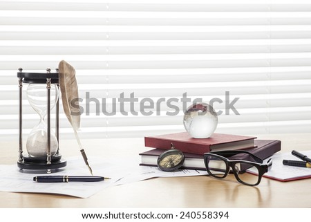 A sand timer(hour glass), feather quill pen, glasses, glass globe, compass, graph paper(document) and book on the wooden office desk(table) behind white blind.