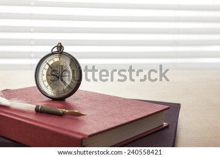A vintage compass, feather quill pen, red book, diary, sketch book on the wooden office desk(table) behind white blind.