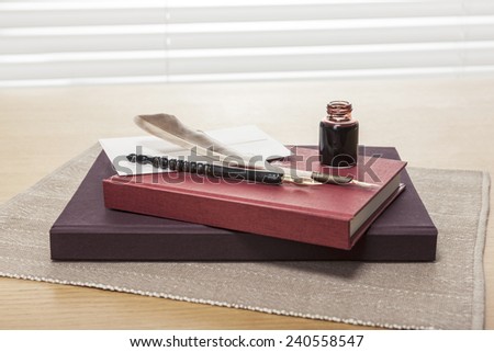envelope, letter, ink, feather quill pen, red book, diary, sketch book on the wooden office desk(table) behind white blind.