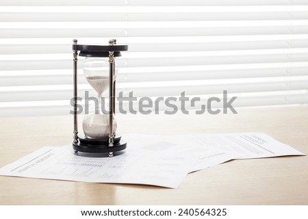 A sand timer(hour glass), graph paper(document) on the wooden office desk(table) behind white blind.