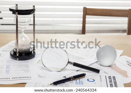 A sand timer(hour glass), glass globe, pen, magnifier, graph paper(document) on the wooden office desk(table) behind white blind.