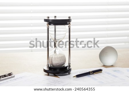 A sand timer(hour glass), glass globe, pen, graph paper(document) on the wooden office desk(table) behind white blind.