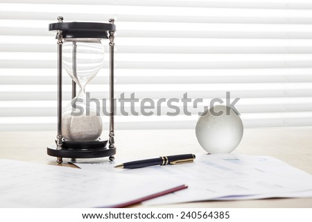 A sand timer(hour glass), glass globe, pen, graph paper(document) on the wooden office desk(table) behind white blind.