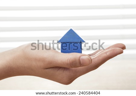 A female(woman) hand hold a blue house(made in wood blocks) white blind background and table.