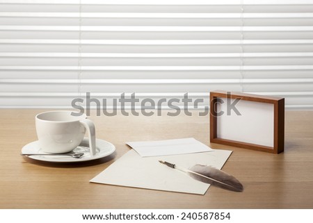 A coffee cup, feather quill pen, wood frame, letter, envelope on the office desk(table) behind white blind.