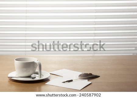 A coffee cup, feather quill pen, letter, envelope on the office desk(table) behind white blind.