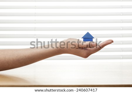 A female(woman) hand hold a blue house(made in wood block) on the office desk(table) behind white blind.