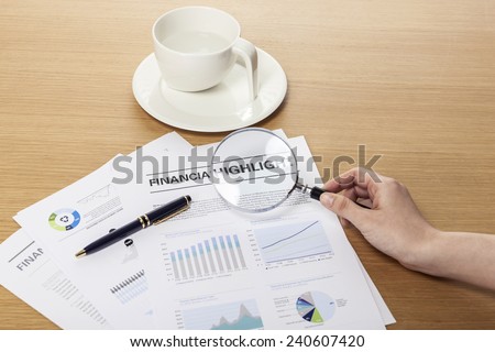 A female(woman) hand hold a magnifier(reading glass) point to graph paper(document), pen on the office desk(table).