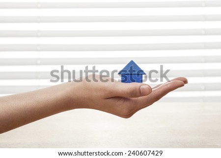 A female(woman) hand hold a blue house(made in wood block) on the office desk(table) behind white blind.