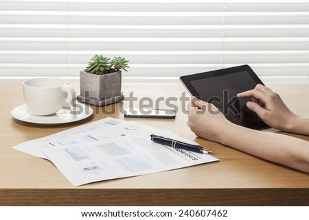 A female(woman) hand hold a tablet pc with graph paper(document) and pen, coffee, mobile phone(hand phone) on the office table(desk) behind white blind.