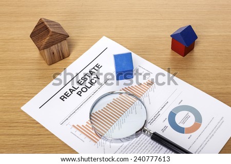 A magnifier and three wood block houses(red, blue) on the graph paper(document) on the wood office desk..