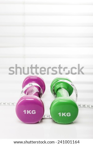 Two dumbbells(pink, green) with tape measure(tape line) on the wood office desk(table) behind white blind.