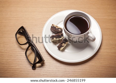 A white coffee cup and saucer with chocolate cookies and black glasses on the wood table(desk)