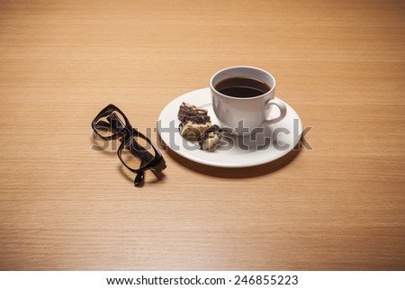 A white coffee cup and saucer with chocolate cookies and black glasses on the wood table(desk)