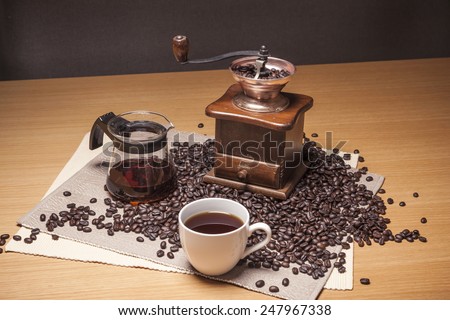 A brass coffee pot, grinder, white cup, beans, table cloth on the wood table(desk).