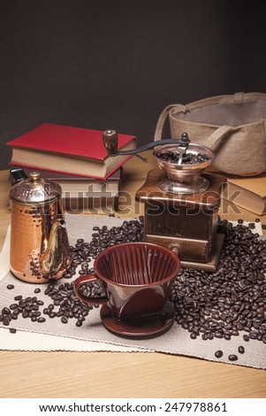 A vintage(old, classic) coffee grinder and books, brass pot, coffee beans, brown dripper on the table cloth and wood table(desk).