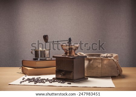 A white coffee cup and wood(vintage, old) grinder, old book, beans, linen bag on the table cloth and wood table(desk).