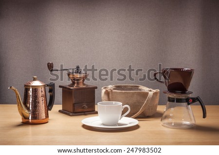 A white coffee cup and wood(vintage, old) grinder, old book, beans, linen bag on the table cloth and wood table(desk).