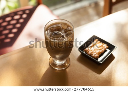 A glass cup of ice black coffee and cookies on the dish on the wood table with shadow of leaves at the early spring, summer.