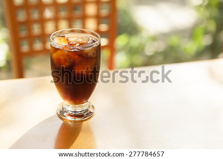 A glass cup of ice black coffee on the wood table with shadow of leaves at the early spring, summer.