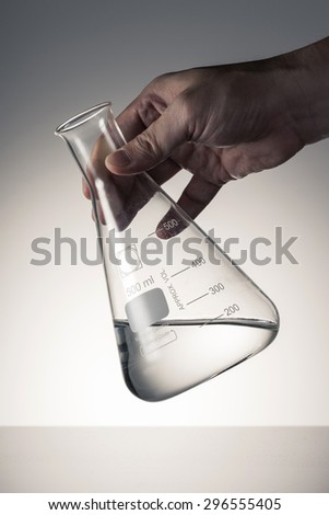 A man\'s hand hold and lean a glass Erlenmeyer flask with water(liquid, fluid) on the reflected desk in the laboratory