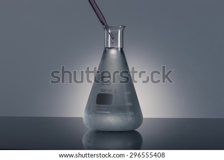A glass Erlenmeyer flask with red(magenta, pink) water(liquid, fluid) and glass dropping pipet(dropping pipette, medicine dropper, spuit) on the reflected black desk in the laboratory