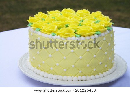 Yellow butter cream frosting handmade roses on a round cake frosted with light yellow icing and embossed with square pattern accented with white frosting dots and white boarder served in back yard