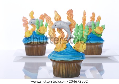 Aquatic under the sea themed cup cakes created at home with royal icing coral and sea weed marshmallow fondant dolphins and fish. Presented on white plates. Perfect for summer parties