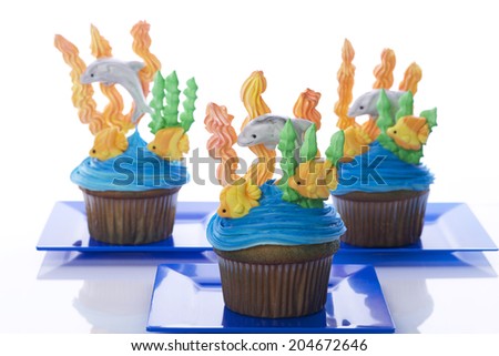 Aquatic under the sea themed cup cakes created at home with royal icing coral and sea weed marshmallow fondant dolphins and fish. Presented on blue plates. Perfect for summer parties