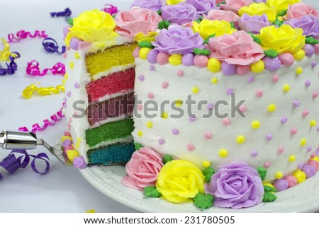Pastel Rainbow Yellow, Pink, Purple butter cream frosting handmade roses on a round cake frosted with white icing with dots of buttercream frosting border. Sliced showing rainbow layered vanilla cake.
