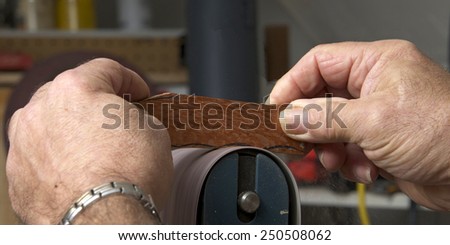 Male hands holding a small piece of cut wood to a table mounted belt sander to smooth the edges on a wood working project. Saw dust everywhere.