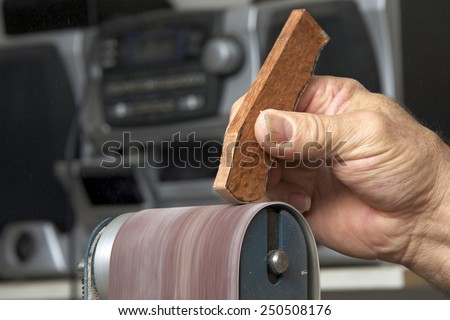 Male hand holding a small piece of cut wood to a table mounted belt sander to smooth the edges on a wood working project. Saw dust everywhere.