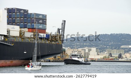 OAKLAND, CA - MARCH 21, 2015: Tugboat AHBRA FRANCO at the Stern of Cargo Ship APHRODITE assisting it to depart the Port of Oakland. A sail boat travels very close to the ship in the Middle Harbor.