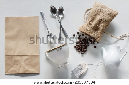 coffee beans in burlap bag, paper bag, silver spoon, vintage cup, white jar, teabag on white wood background.