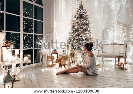 woman in white sweater, warm and cozy evening in Christmas interior design, Xmas tree decorated by lights gifts toys, candles, lanterns, garland lighting indoors fireplace.holiday living room.New year