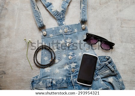 Outfit of casual woman. Blue denim overall mobile phone sunglasses leather bracelet on a wooden background