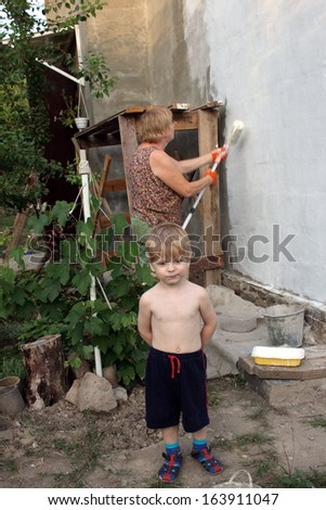 small boy posing on the backdrop of middle-aged woman paints his own house a paint roller on a long handle