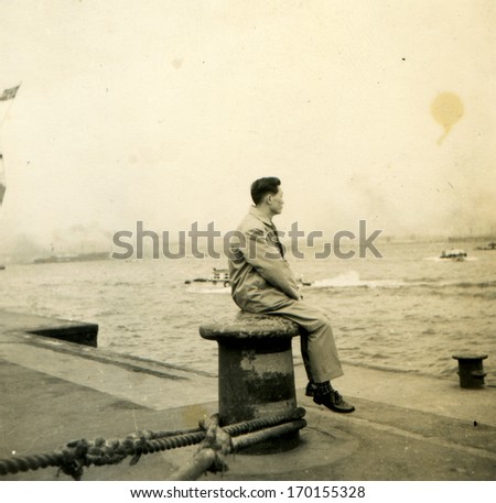 JAPAN  - Circa 1950s: An antique photo shows man sitting on a bollard on the dock watching the sea