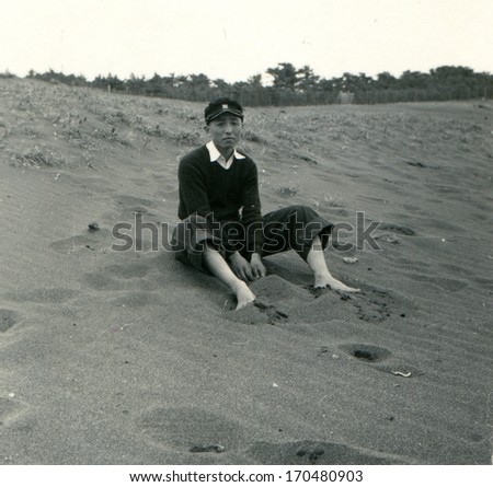 JAPAN  - 1950s: An antique photo shows young man in a white shirt, a dark sweater and cap student sits on a sand hill